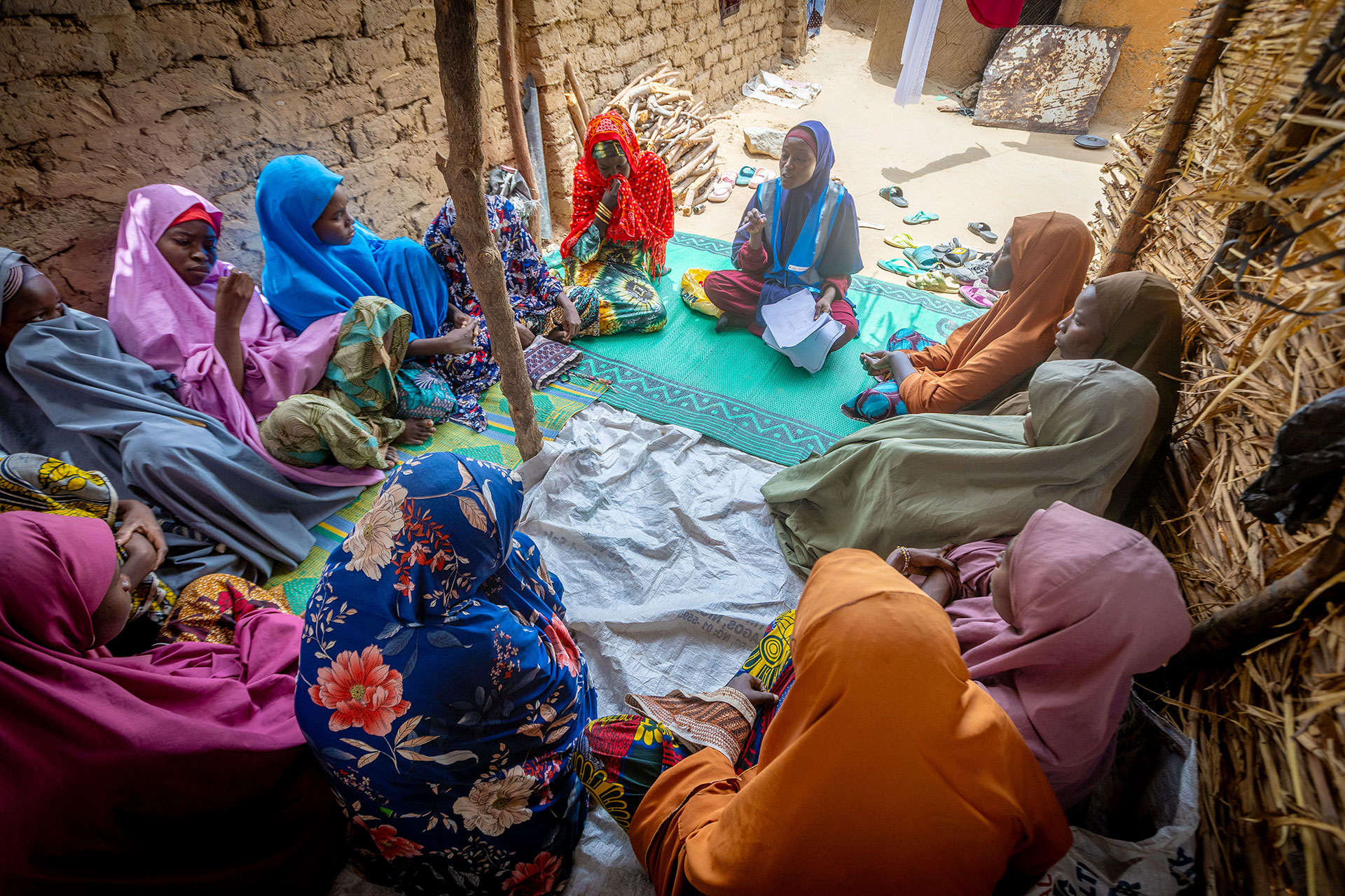A group of women wearing colorful headscarves and dresses sit in a circle on mats in an outdoor, narrow, clay-walled alley, engaged in conversation or discussion. Sandals are scattered on the ground nearby. A group of women wearing colorful headscarves and dresses sit in a circle on mats in an outdoor, narrow, clay-walled alley, engaged in conversation or discussion. Sandals are scattered on the ground nearby.