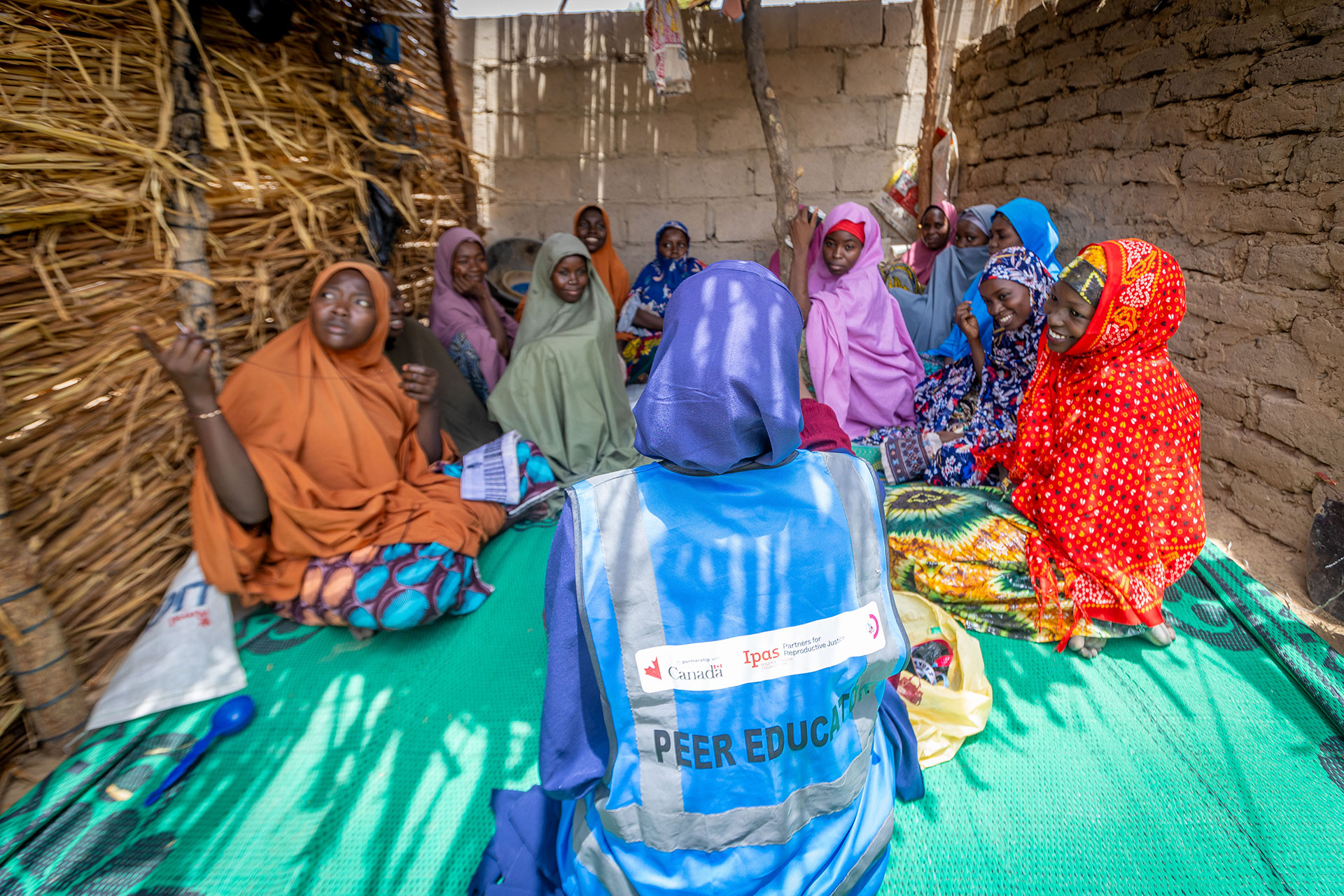 A group of women in colorful clothing sit on a mat inside a thatched and mud-walled shelter, listening to a person wearing a blue vest labeled "PEER EDUCATOR" at the front. A group of women in colorful clothing sit on a mat inside a thatched and mud-walled shelter, listening to a person wearing a blue vest labeled "PEER EDUCATOR" at the front.