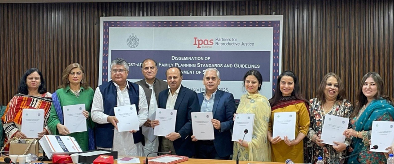 A group of men and women stand together indoors, holding documents and smiling at the camera. Behind them is a banner that reads, "Dissemination of Post-Abortion Family Planning Standards and Guidelines with the Ipas Pakistan logo at top.