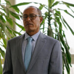 A man wearing glasses and a gray suit with a light blue tie stands indoors in front of large green plants and a window.