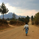 A person wearing a white shirt and blue patterned skirt walks alone on a wide, dusty road in Malawi, lined with dry bushes and trees. In the distance, mountains rise beneath a cloudy sky—symbolic of abortion access survivors' journeys.