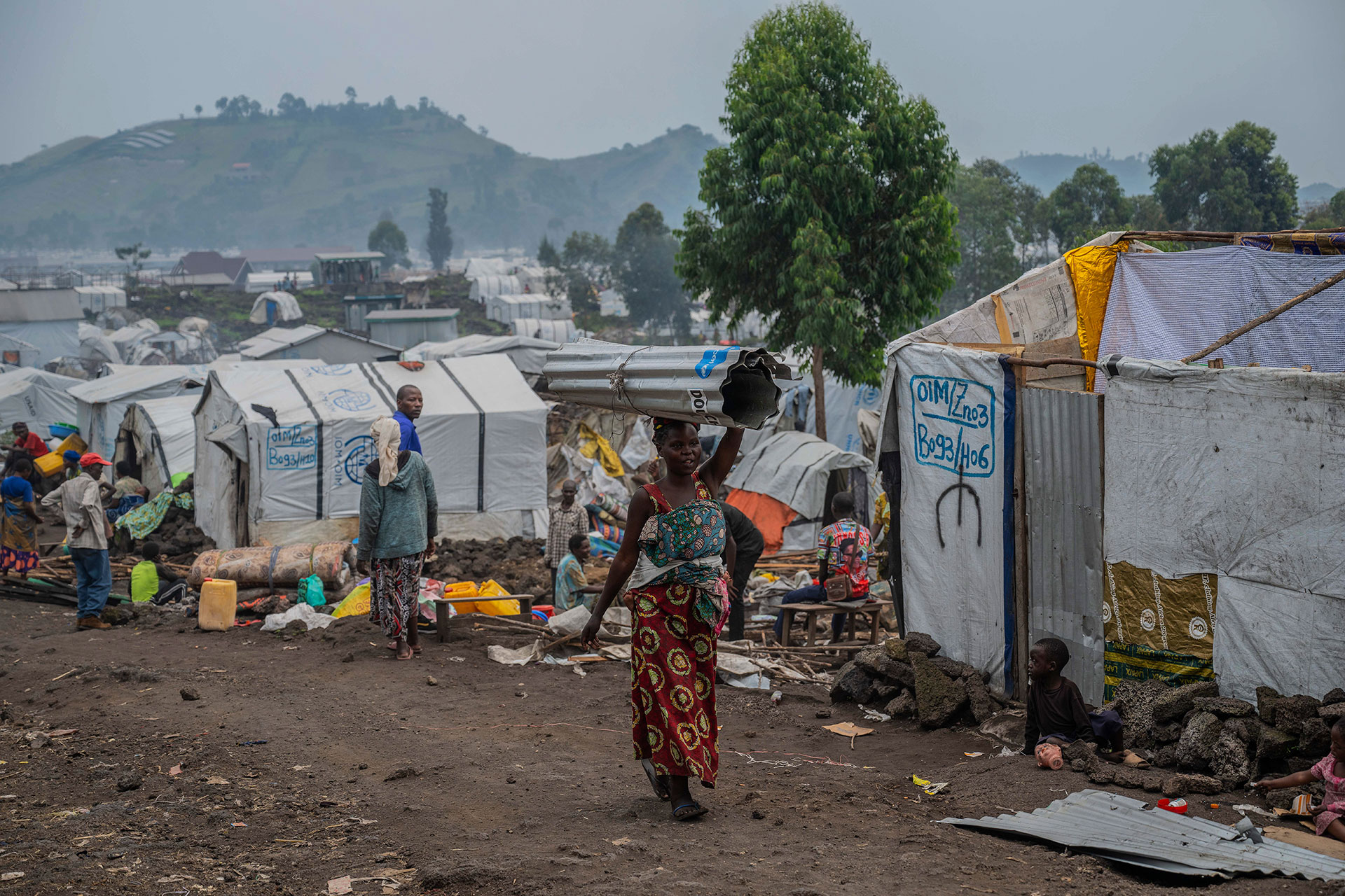 A woman carries a metal sheet on her head in a camp with makeshift tents, surrounded by scattered belongings and other people, with hills and trees in the background.