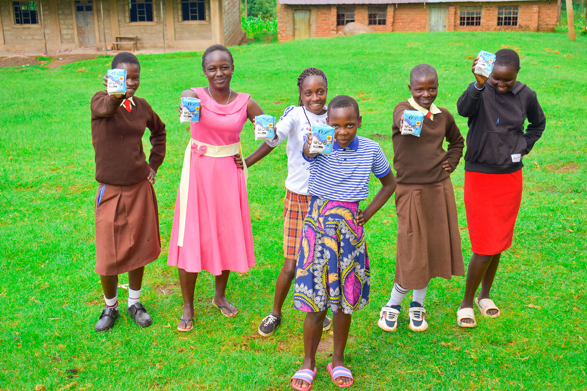 Six smiling girls stand outside on green grass, holding up packaged items toward the camera. They wear colorful skirts, dresses, and school uniforms, with buildings visible in the background.