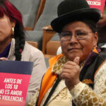 Two women sit in an auditorium holding a sign that reads “Antes de los 18 no es amor, es violencia!” (“Before 18, it’s not love, it’s violence!”). One woman gives a thumbs-up gesture.