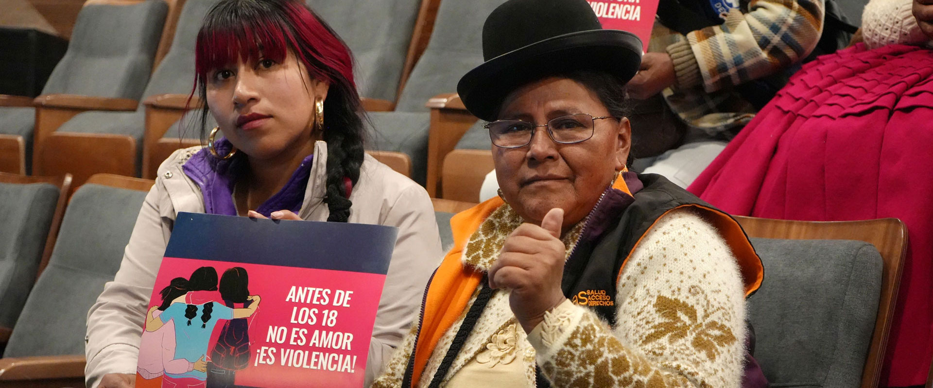 Bolivia Chamber of Deputies Two women sit in an auditorium holding a sign that reads “Antes de los 18 no es amor, es violencia!” (“Before 18, it’s not love, it’s violence!”). One woman gives a thumbs-up gesture.