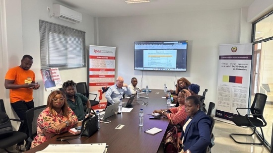 A group of people sit around a conference table with laptops and documents, attending a meeting in a modern office. Posters and banners with text are displayed on the walls, and a screen shows a document.