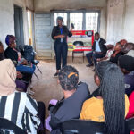 A group of people sits in a circle on black chairs inside a plain, unfinished room, while a person standing at the front speaks to them. Light comes in through windows at the back.