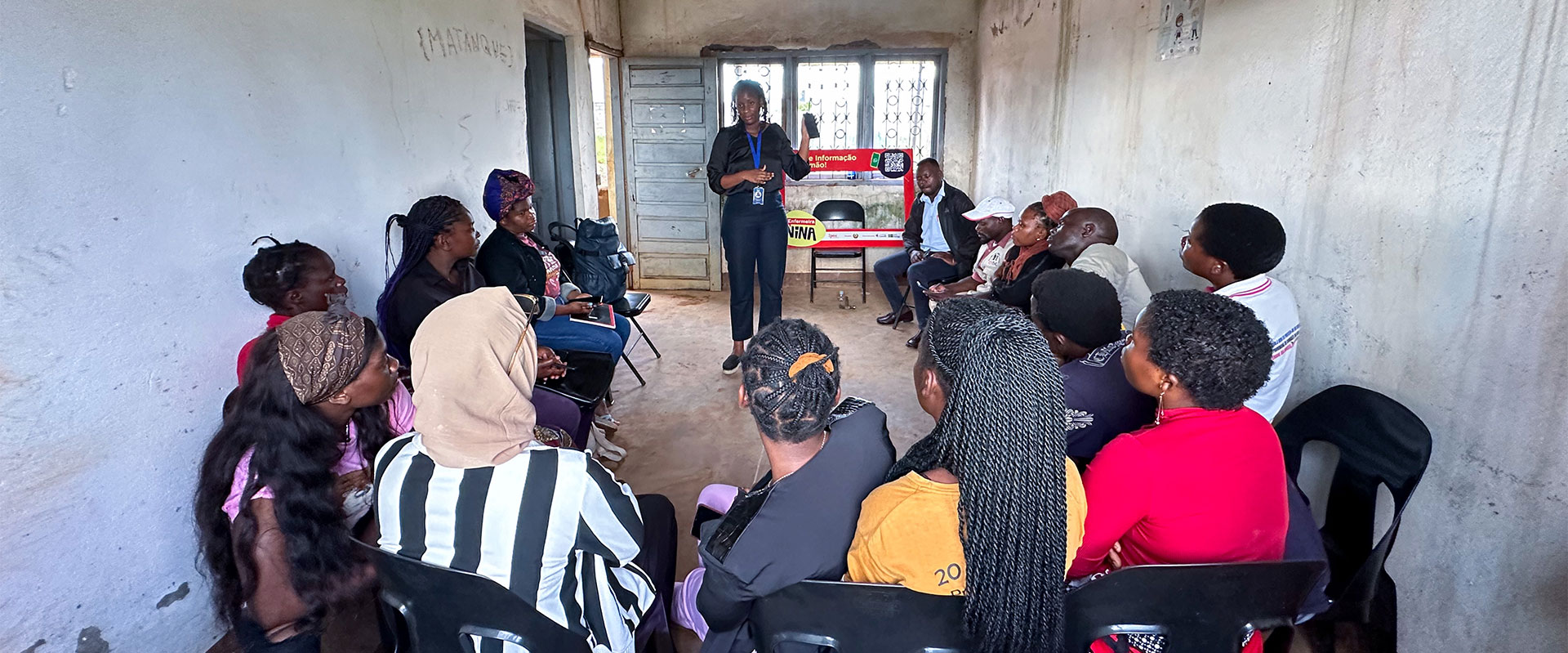 A group of people sits in a circle on black chairs inside a plain, unfinished room, while a person standing at the front speaks to them. Light comes in through windows at the back.