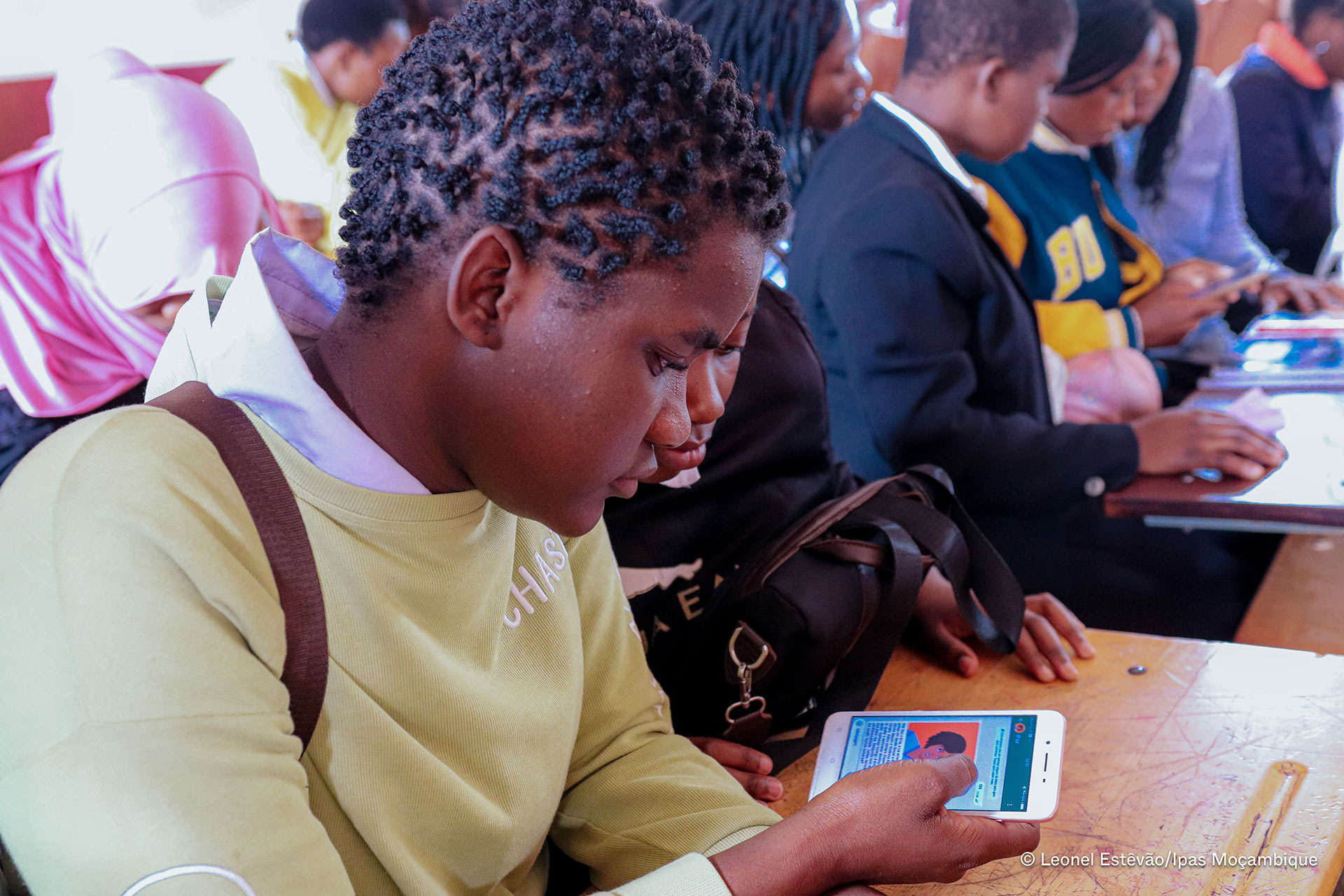 A student in a classroom intently uses a smartphone, with other students also engaged with their devices in the background. The scene suggests a technology-based learning environment.