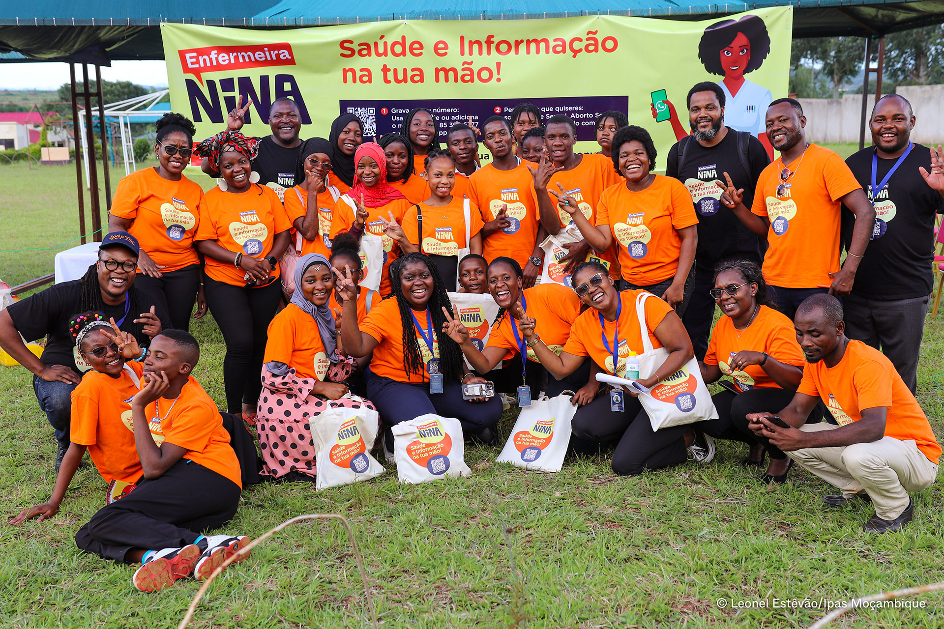 A group of smiling people in orange shirts pose together outdoors in front of a banner that reads “Enfermeira NENA” and “Saúde e Informação na tua mão!” Some hold tote bags and make peace signs.