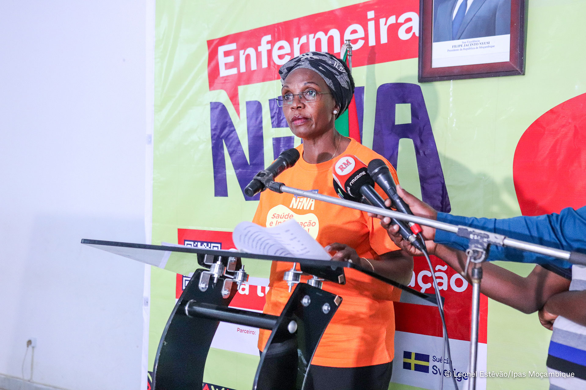 Governor Niassa, Elina Judite Massengele , a woman wearing glasses and an orange shirt speaks at a podium with microphones. Behind her is a green and red banner with Portuguese text and a framed photo of a man on the wall.