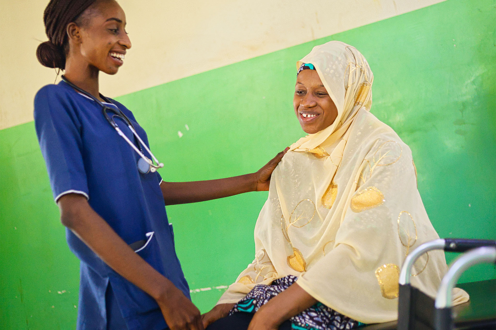 A smiling healthcare worker in blue scrubs chats with a seated woman in a cream hijab and patterned dress, gently resting a hand on her shoulder, against a green and beige wall.