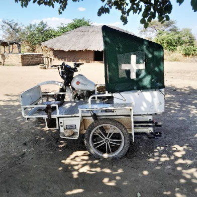 A three-wheeled motorcycle ambulance with a covered compartment displaying a white medical cross is parked on dirt ground near traditional thatched-roof huts, with trees and blue sky in the background.