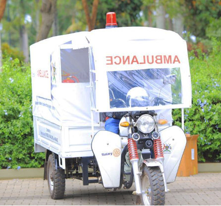 A three-wheeled motorcycle ambulance with a white cover and a red siren on top is parked on a paved path, surrounded by greenery. The driver, wearing a helmet, is partially visible inside.