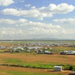 A wide view of a refugee camp with many white tents and small structures scattered across a dry, open landscape under a partly cloudy sky, with mountains visible in the distance.