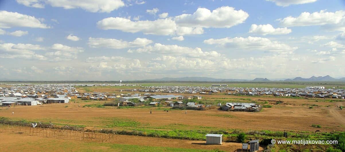 A wide view of a refugee camp with many white tents and small structures scattered across a dry, open landscape under a partly cloudy sky, with mountains visible in the distance. A wide view of a refugee camp with many white tents and small structures scattered across a dry, open landscape under a partly cloudy sky, with mountains visible in the distance.
