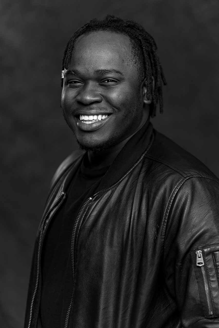 Portrait of Kondwani Jere who is smiling, with short dreadlocks wearing a black leather jacket and a dark shirt, photographed against a dark, blurred background. The image is in black and white.