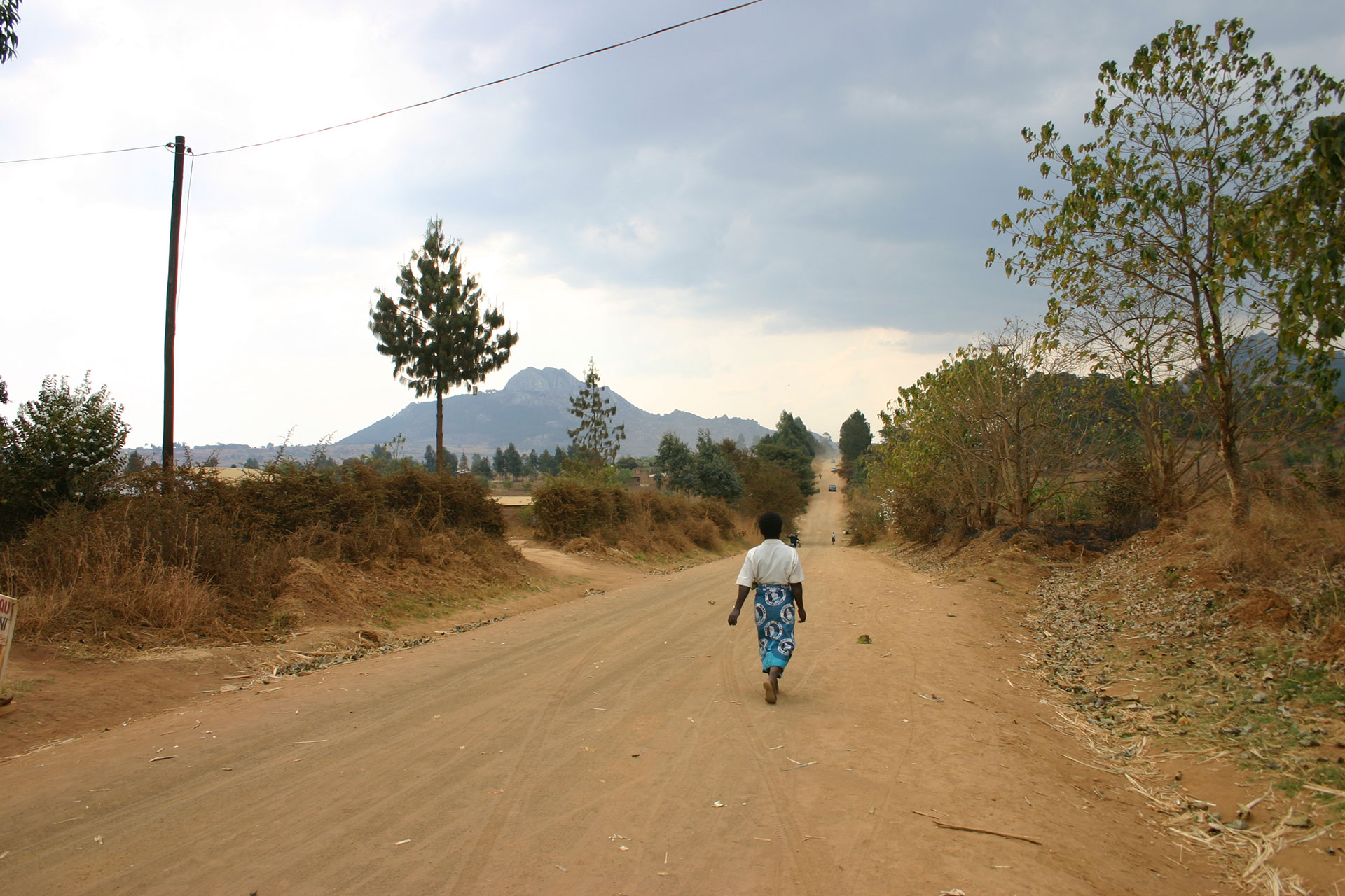 A person wearing a white shirt and blue patterned skirt walks alone on a dusty rural road lined with dry trees and bushes, with mountains visible in the distance under a cloudy sky.