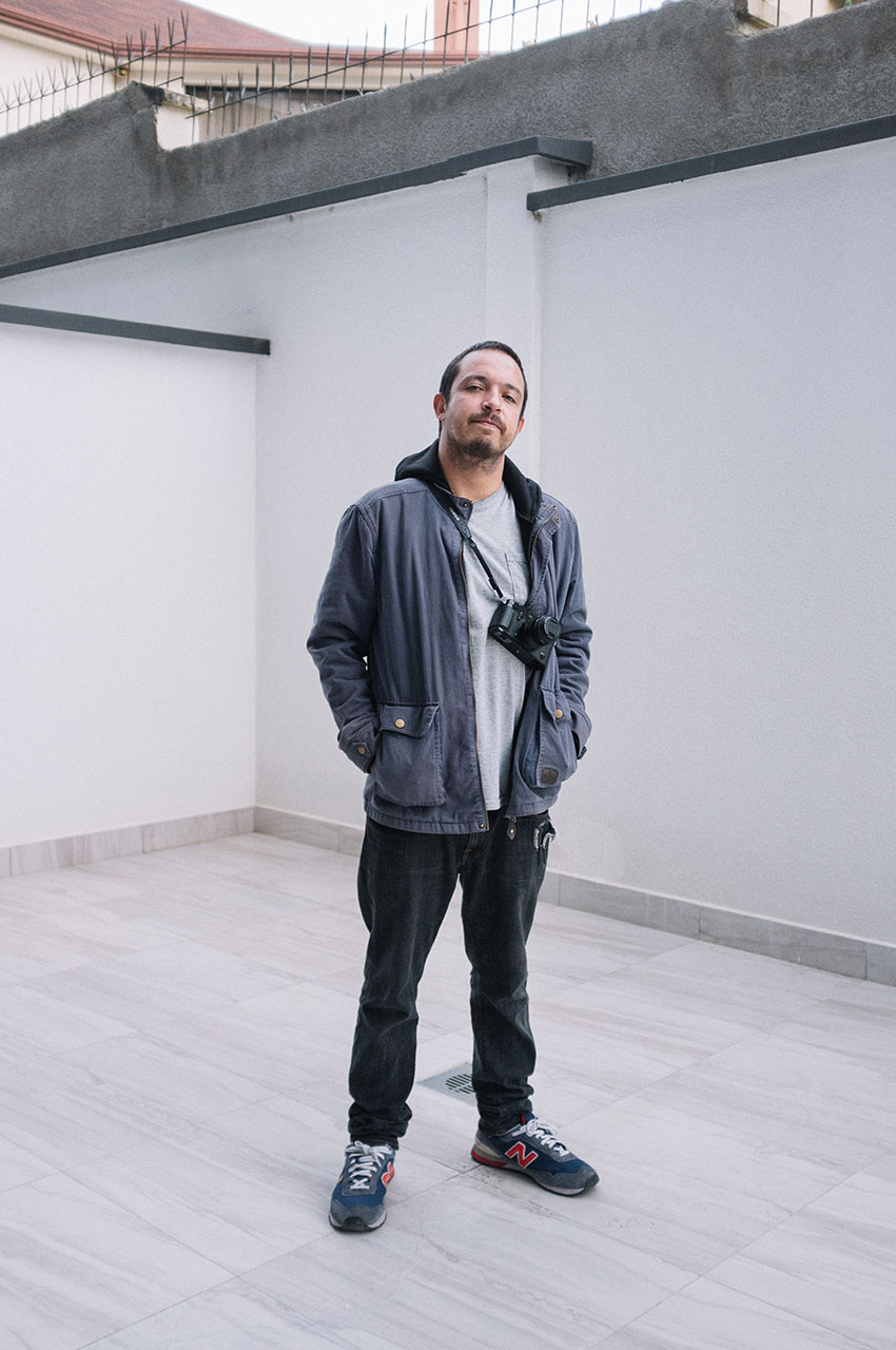 Portrait of Manuel Seoane, who has a short beard standing in a minimal, empty outdoor space. He wears a blue jacket, gray shirt, dark jeans, sneakers, and has a camera strap around his neck. He looks at the camera with a relaxed posture.