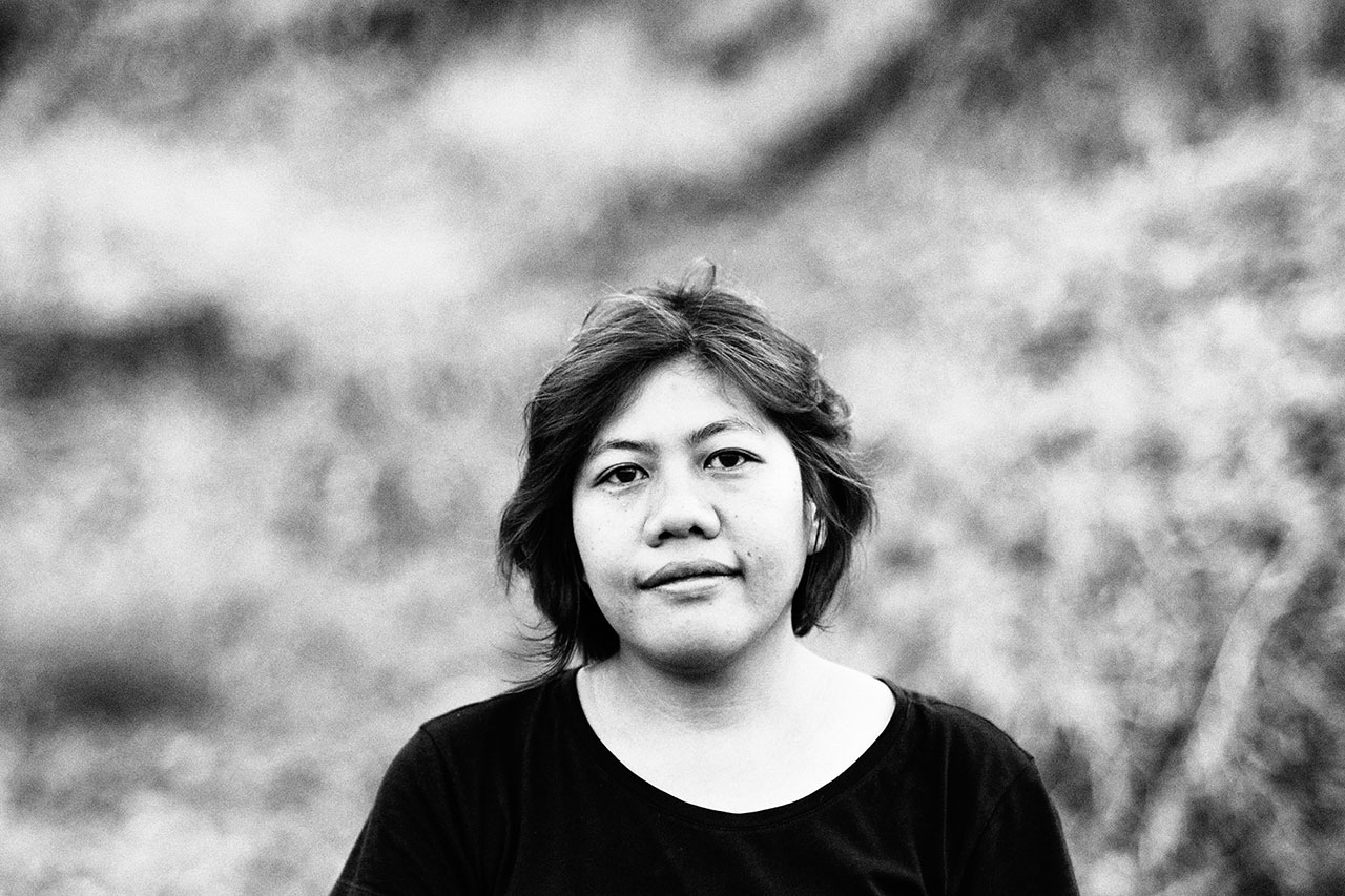 Black and white portrait of Rosa Panggabean who has short hair wearing a dark shirt, looking directly at the camera with a neutral expression; blurred outdoor background.