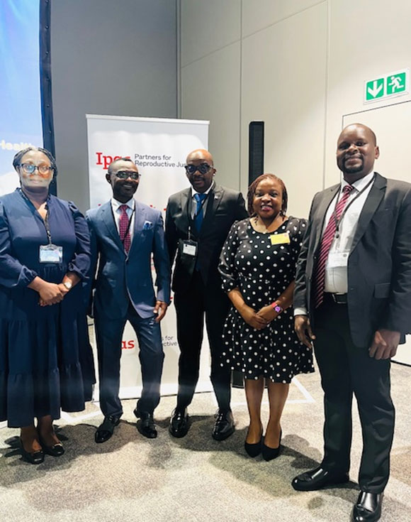 Five people wearing business attire stand indoors in front of an IPPF banner and a white wall. Three men and two women smile for the photo.