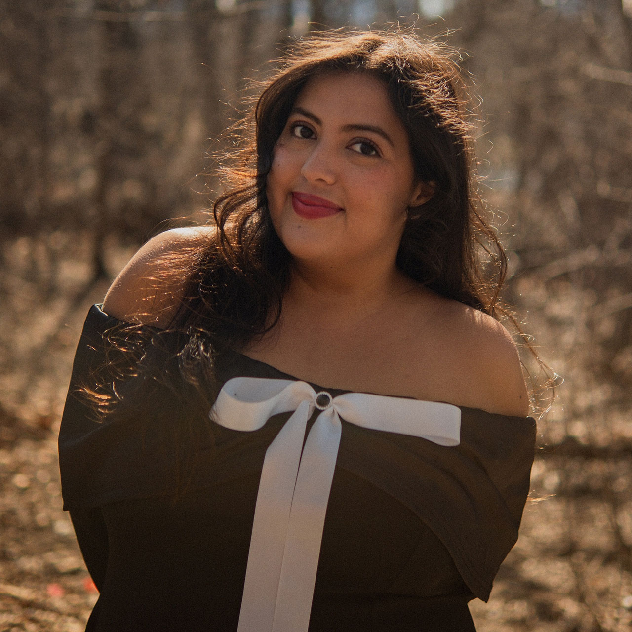 A woman with long dark hair, wearing an off-the-shoulder black dress with a large white bow, smiles outdoors in a sunlit, leafless forest.
