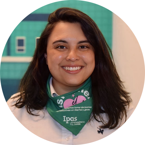 A person with long dark hair smiles at the camera, wearing a white shirt and a green bandana with text and logos around their neck. The background is teal with some blurred shapes.