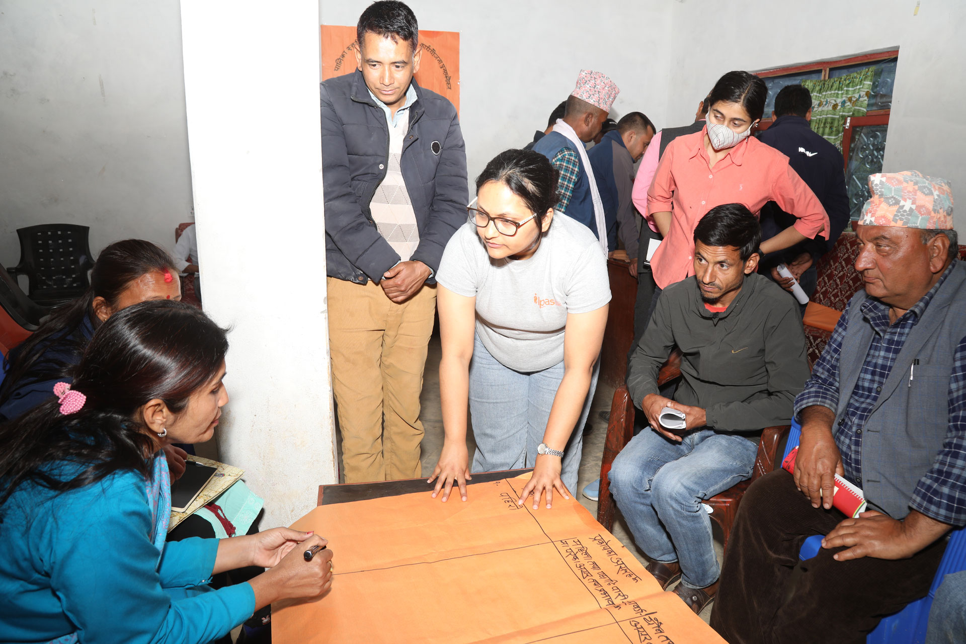 A group of people sit and stand around a table with large sheets of paper, discussing and writing notes in a brightly lit room. Some participants are actively engaged, while others observe.
