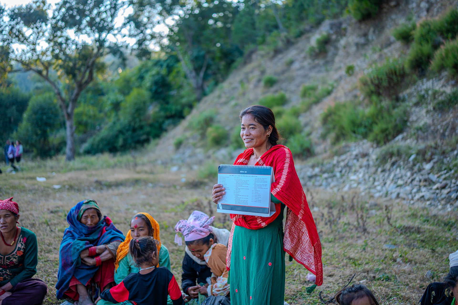 A woman stands outdoors holding up a paper with printed text, smiling at a small group of seated people. The group is on grassy ground near a hillside, with trees in the background.