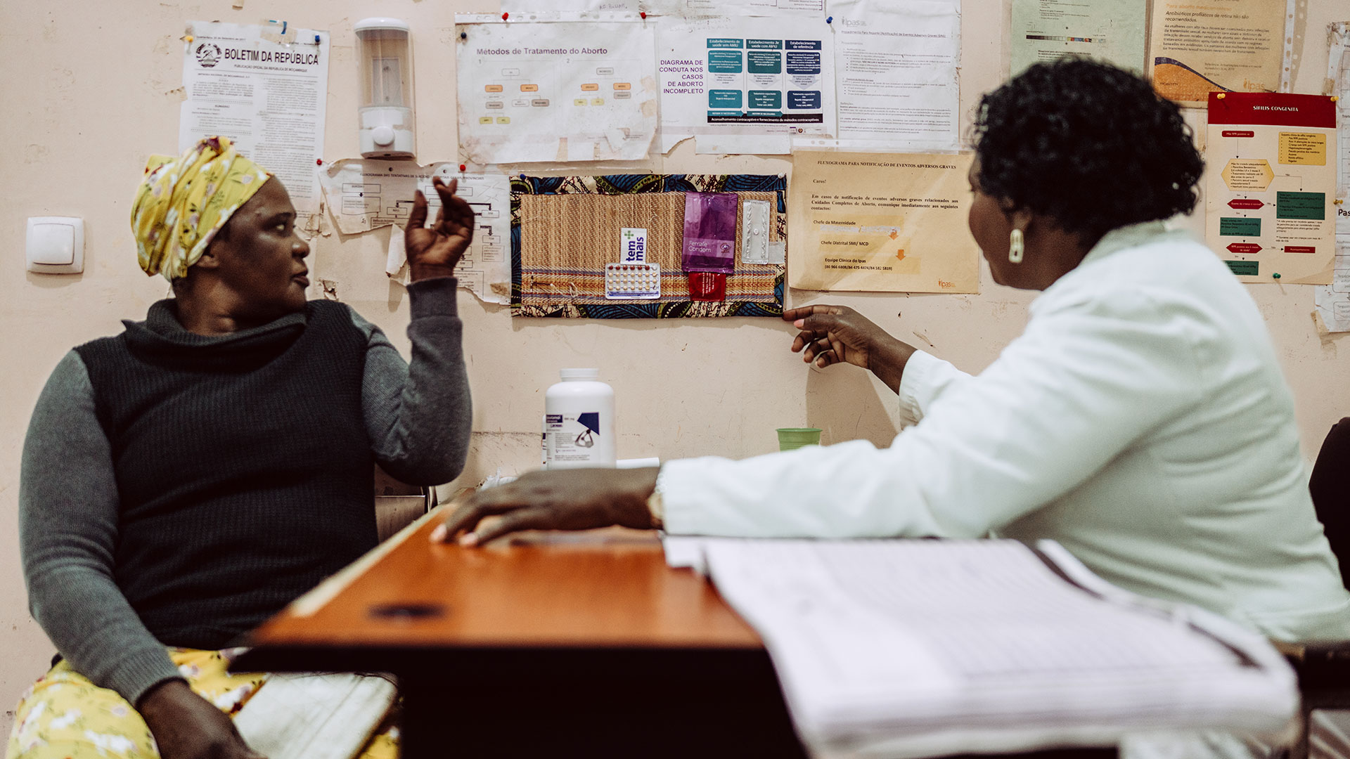 Two women sit across from each other at a desk in a medical office, discussing information on a wall covered with charts and papers. One woman is dressed in a white coat, the other wears a headscarf and sweater.