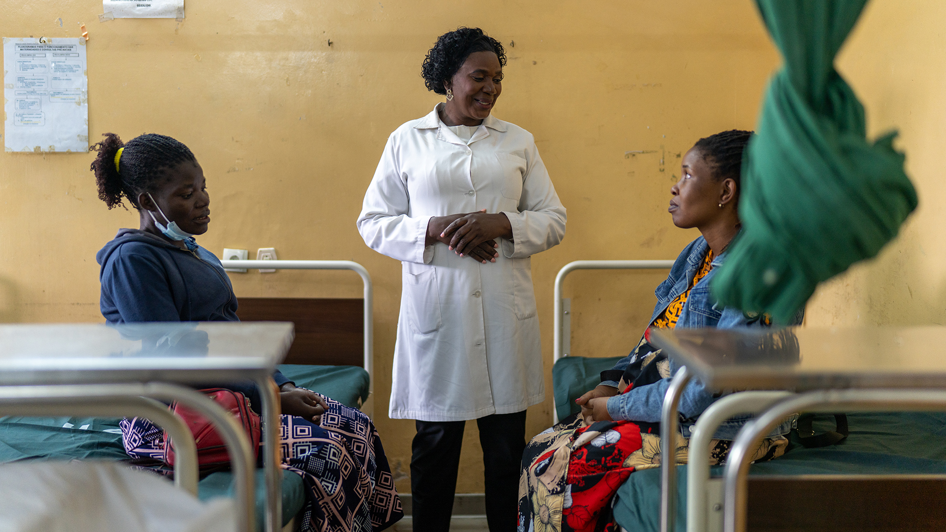 A healthcare worker in a white coat stands between two women sitting on hospital beds, engaging in conversation in a medical ward with yellow walls.