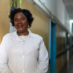 Estrela Góia, a woman in a white lab coat stands smiling against a yellow wall in a well-lit hospital hallway, with blue doors visible along the corridor.