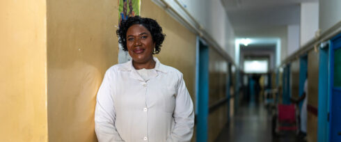 Estrela Góia, a woman in a white lab coat stands smiling against a yellow wall in a well-lit hospital hallway, with blue doors visible along the corridor.