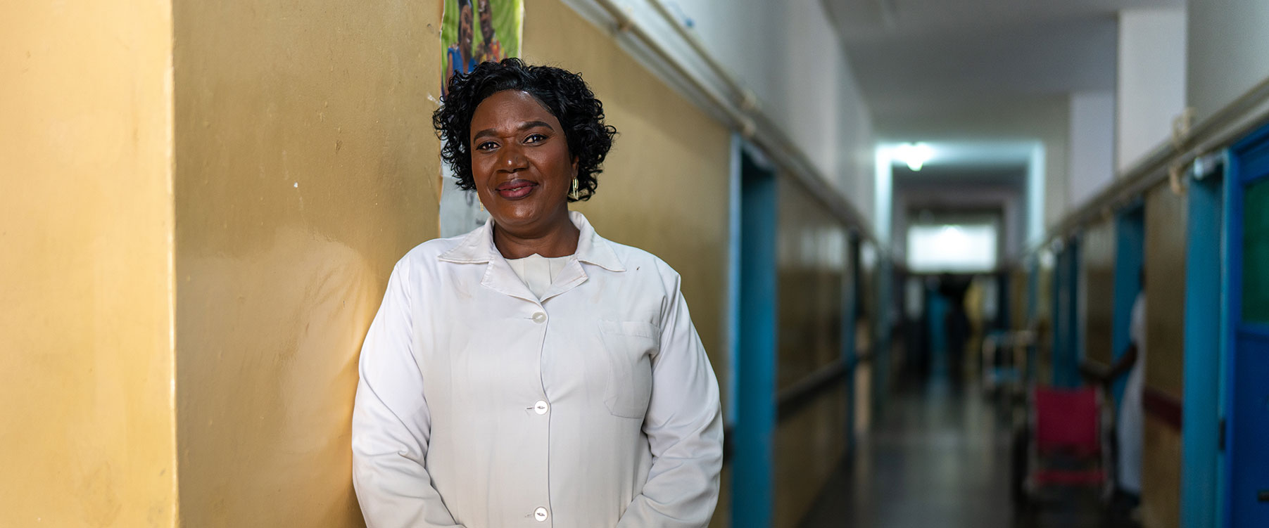 Estrela Góia, a woman in a white lab coat stands smiling against a yellow wall in a well-lit hospital hallway, with blue doors visible along the corridor.