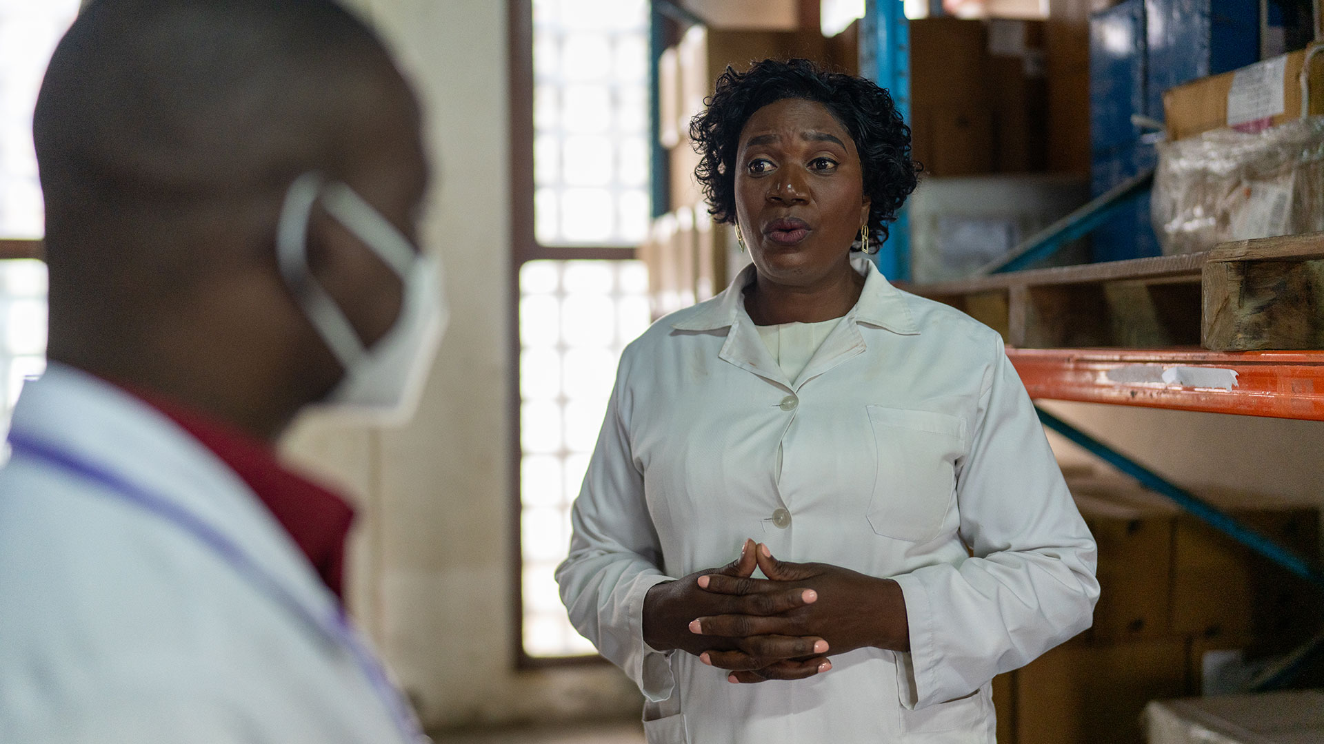 A woman in a white lab coat speaks to a colleague wearing a face mask in what appears to be a medical or laboratory storage room with shelves and boxes.