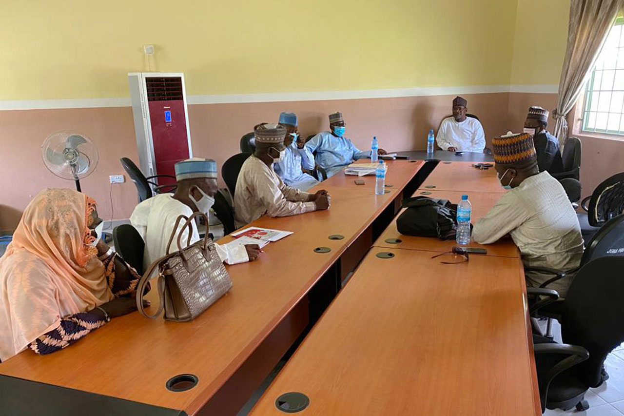 Several people sit around a large conference table in a meeting room, with notebooks, water bottles, and bags on the table. Most attendees wear traditional clothing and hats, and sunlight enters through a window.