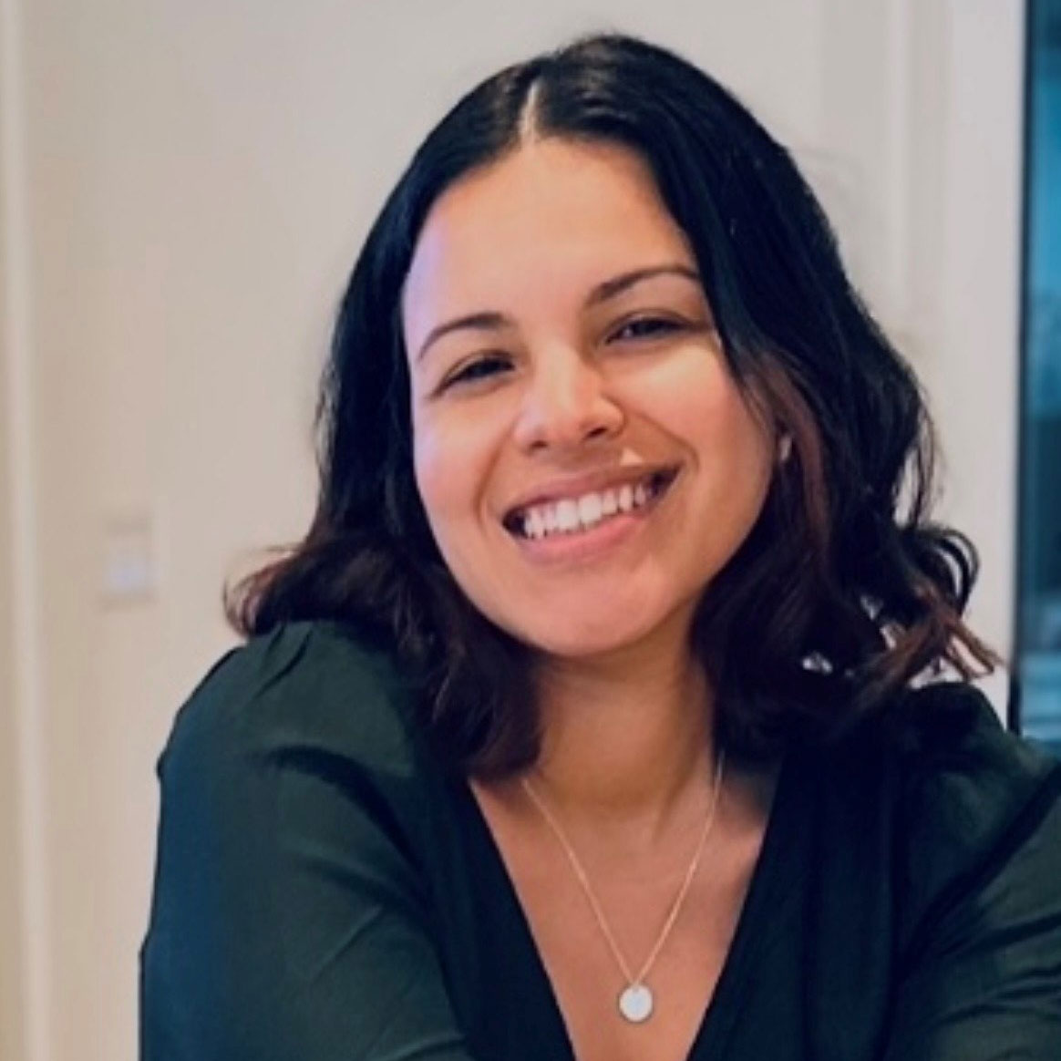 A woman with shoulder-length dark hair smiles warmly at the camera. She is wearing a dark top and a necklace with a round pendant, sitting indoors with a light background.