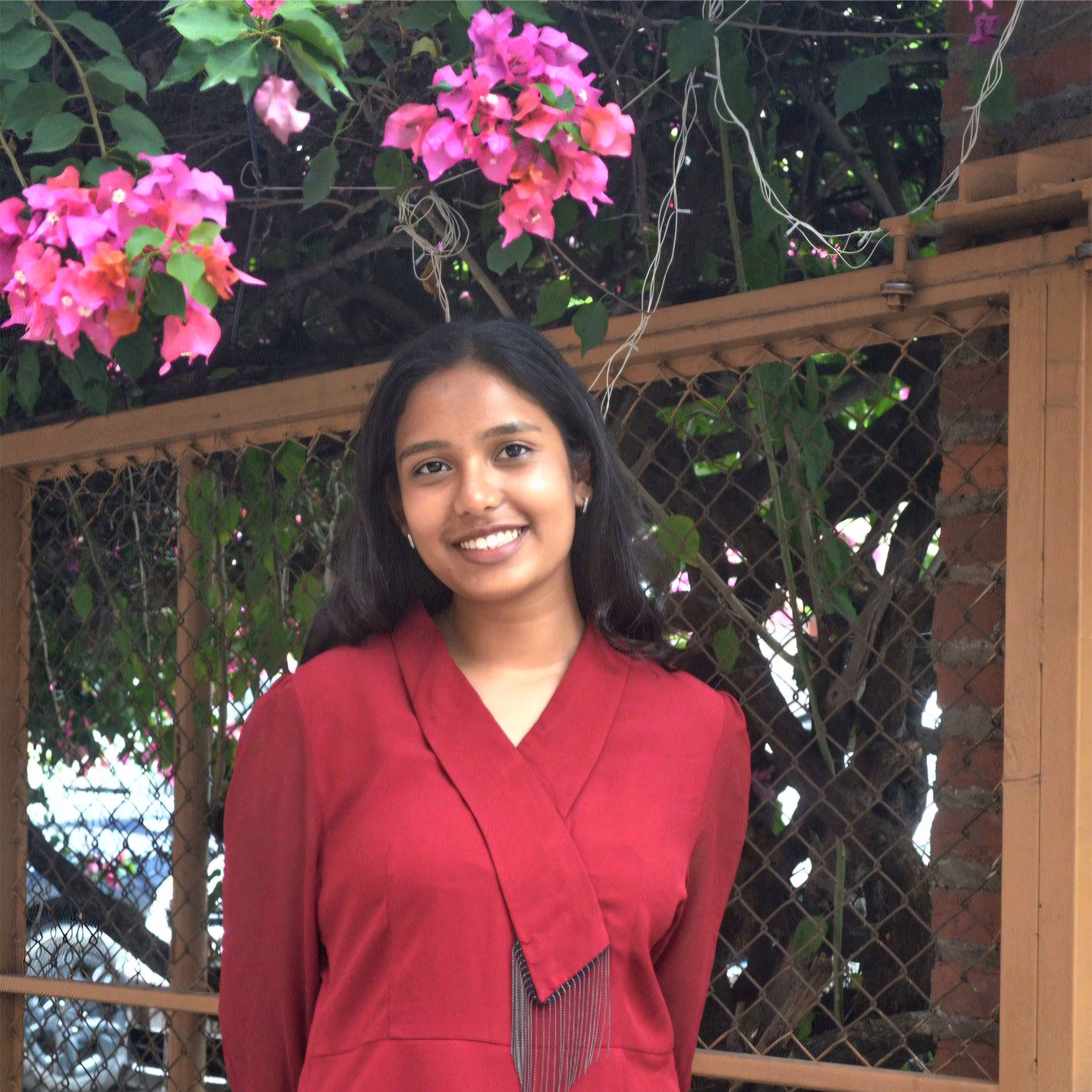A young woman in a red dress smiles while standing in front of a wooden fence with blooming pink flowers and green foliage above her.