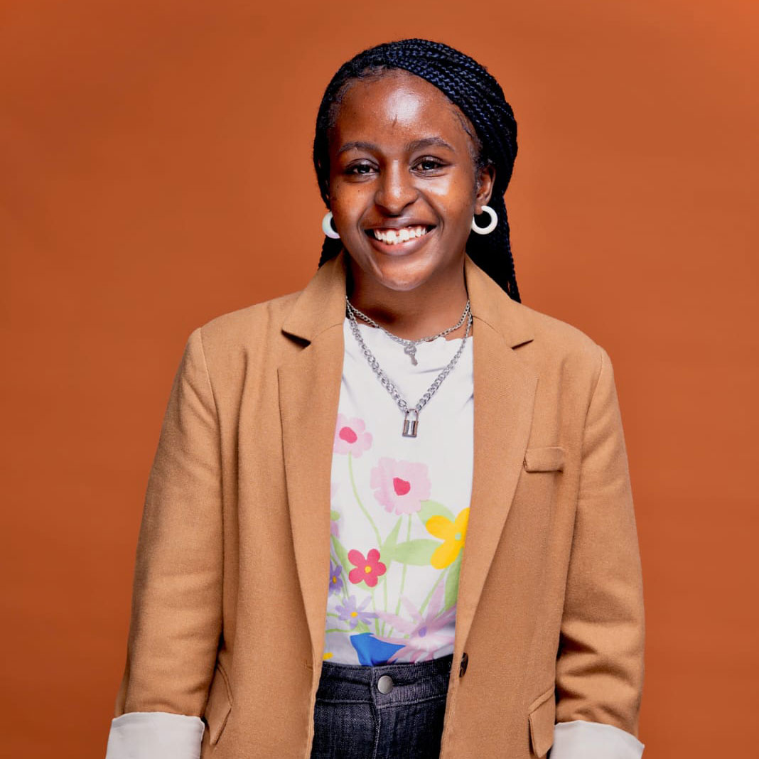 A smiling person with braided hair wears hoop earrings, layered necklaces, a tan blazer over a floral shirt, and dark pants, standing against a solid orange background.