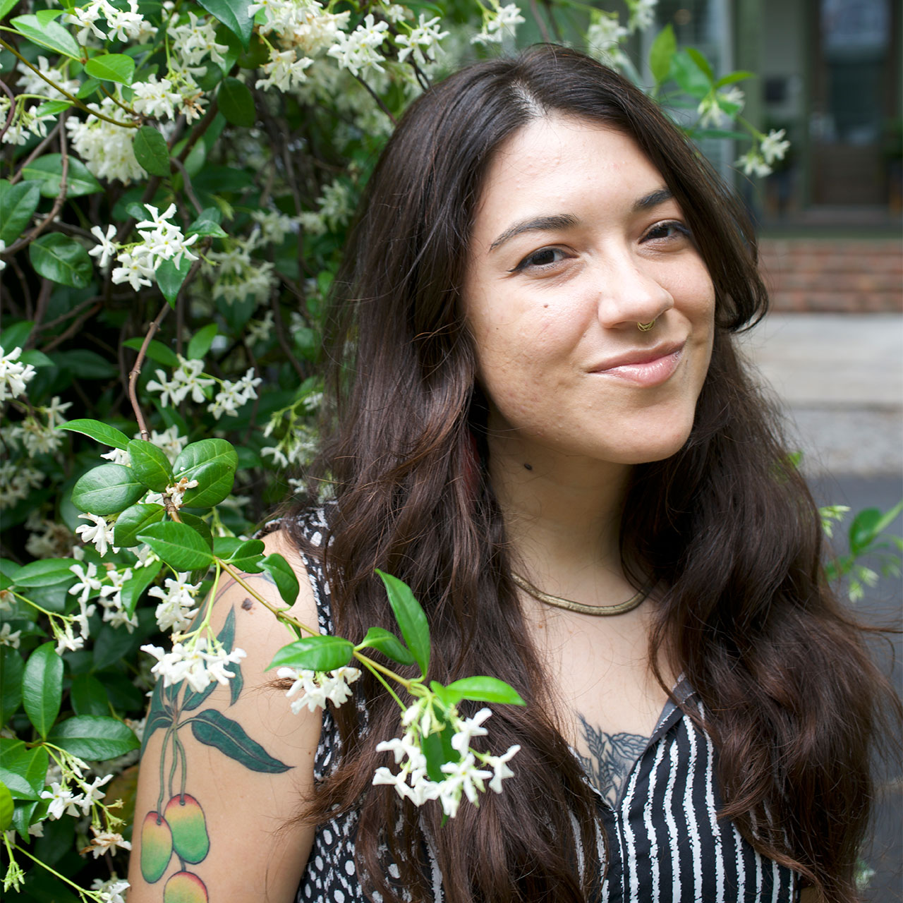A woman with long brown hair and a nose ring smiles slightly while standing in front of a bush with white flowers. She is wearing a striped sleeveless top and has a colorful fruit tattoo on her arm.