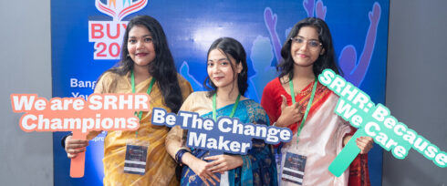 Three women in traditional attire hold colorful signs reading “We are SRHR Champions,” “Be The Change Maker,” and “SRHR: Because We Care” at a youth event with a blue backdrop.