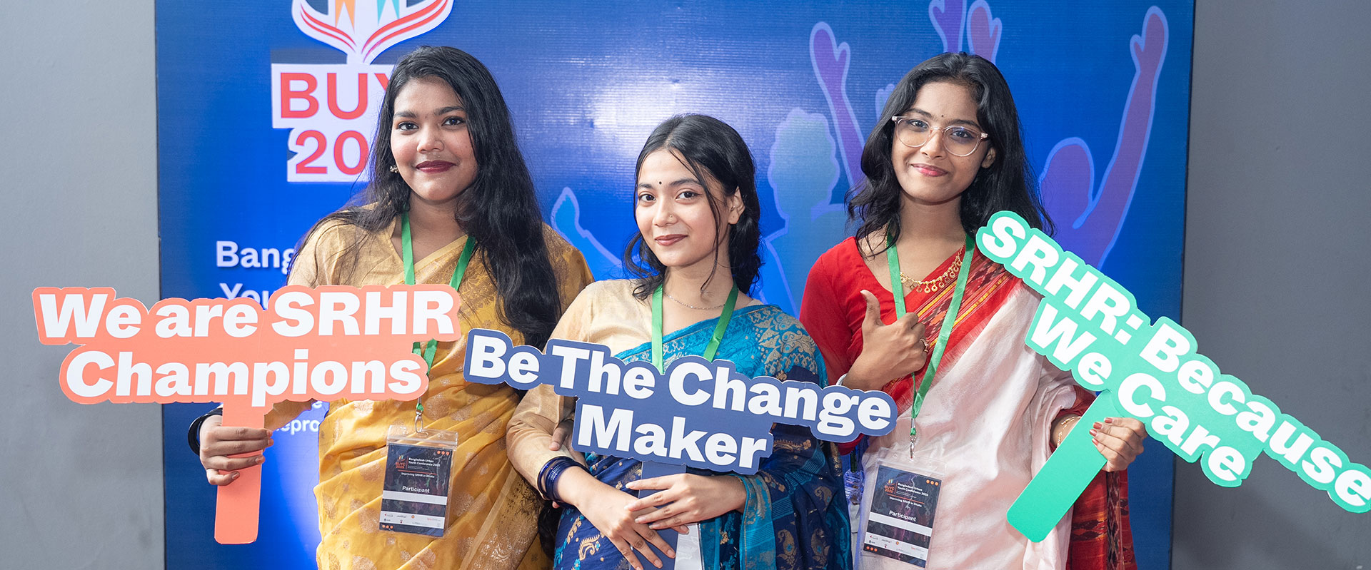 Three women in traditional attire hold colorful signs reading “We are SRHR Champions,” “Be The Change Maker,” and “SRHR: Because We Care” at a youth event with a blue backdrop.
