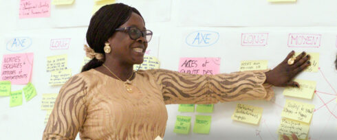 A woman wearing glasses and a patterned dress gestures toward a wall covered with colorful sticky notes and diagrams, appearing to present or explain information.