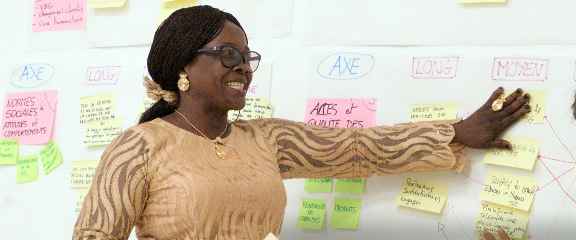 A woman wearing glasses and a patterned dress gestures toward a wall covered with colorful sticky notes and diagrams, appearing to present or explain information.