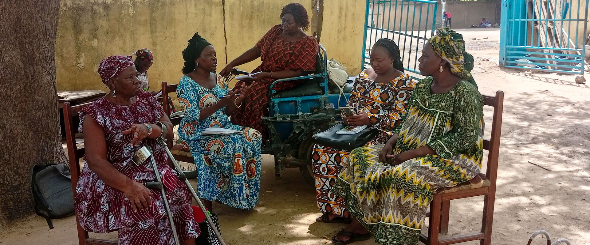 Five women in colorful patterned dresses sit together outside, talking and gesturing. They appear relaxed and engaged in conversation, with a wall and blue metal gate visible in the background.