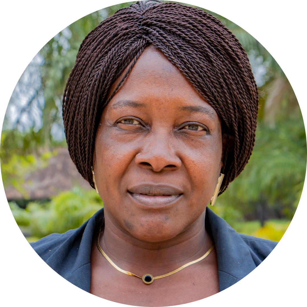 A woman with braided hair looks directly at the camera, wearing a black top, a gold necklace, and gold earrings. Greenery and trees are visible in the blurred background.