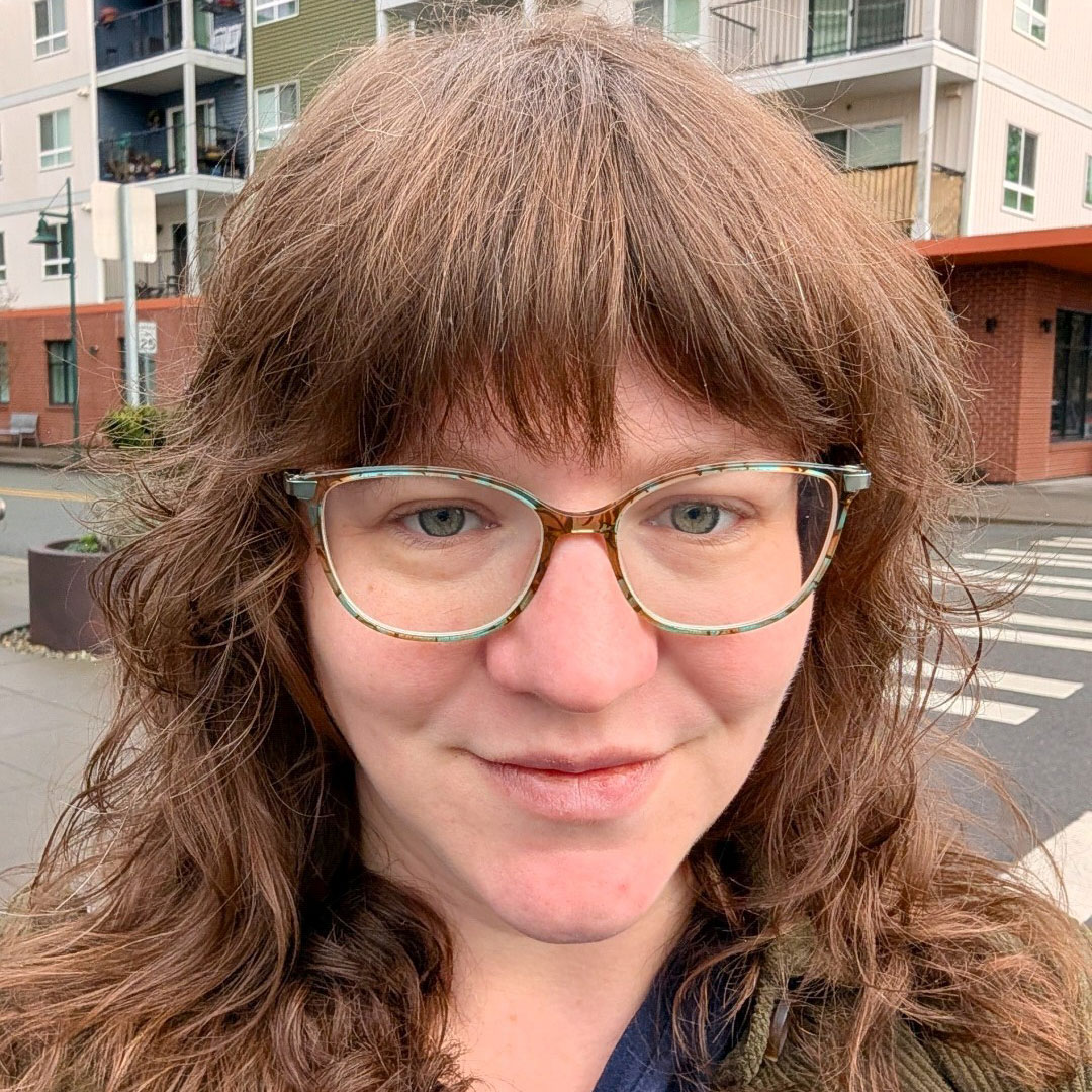 A person with wavy brown hair and light blue glasses smiles outdoors in front of a modern apartment building and crosswalk.