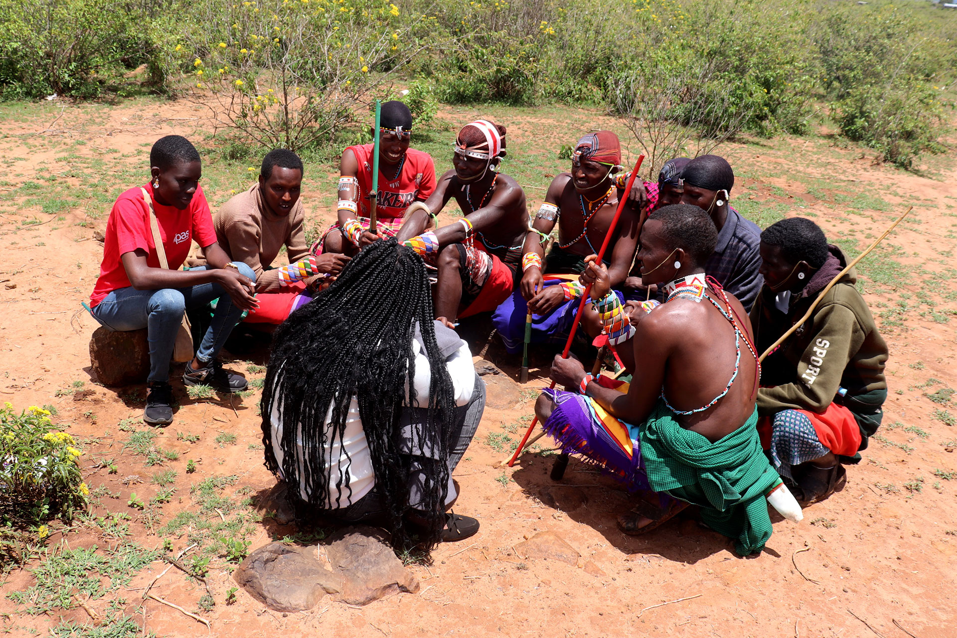 A group of people, some in traditional Maasai clothing and beadwork, sit in a circle outdoors on reddish soil, surrounded by green shrubs. They appear engaged in conversation or an activity.