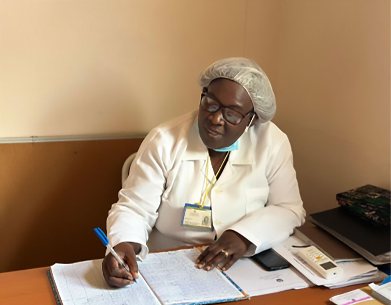 A person wearing a white lab coat, hairnet, and glasses sits at a desk, writing in a large notebook. Office supplies and documents are on the desk. The person has an ID badge and a face mask under their chin.