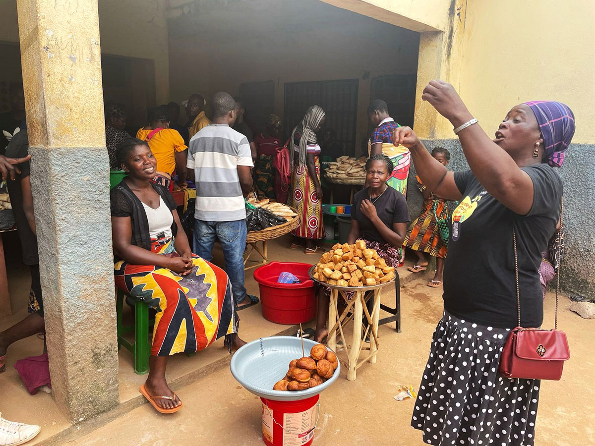 A lively market scene shows women selling and displaying fried snacks on tables. Shoppers and vendors interact, some seated and some standing, with food items in bowls. The setting is a shaded area with yellow walls.